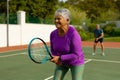 Smiling biracial senior woman with short hair playing tennis in court with senior man in background Royalty Free Stock Photo
