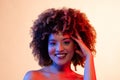 Smiling biracial woman with curly hair in eyeshadow and lipstick touching head in red and blue light Royalty Free Stock Photo
