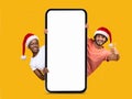 Smiling black and arab millennial woman and guy in Santa Claus hats showing thumb up with big smartphone