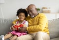 Smiling Black Elderly Man Gives Birthday Present, Little Child Opens Box In Living Room Interior