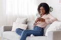 Smiling black girl messaging on smartphone at home, relaxing on comfortable couch Royalty Free Stock Photo