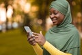 Smiling Black Islamic Woman In Hijab Using Smartphone Outdoors, Browsing Internet