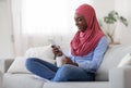 Smiling black muslim lady resting at home, using smartphone and drinking coffee