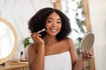 Smiling black woman applying cosmetic powder or blusher on her face, using decorative makeup, looking in mirror at home Royalty Free Stock Photo