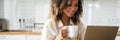 A smiling black woman in a wired headphones with a cup in her hands sitting in front of laptop in the kitchen