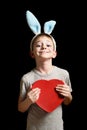 Smiling blond boy in hare ears holds heart shape red box on black background. Love and family concept. Vertical frame