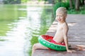 Smiling blond boy sits on a pier near the lake with a circle, swims, a child learns to swim, a child`s rest in the village