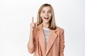 Smiling blond girl in business suit, pointing finger up, looking amazed, standing over white background Royalty Free Stock Photo