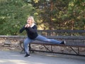 Smiling blond grownup woman in hoodie doing one leg squat using a bench in urban park in summer, outdoor, selective Royalty Free Stock Photo