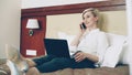 Smiling businesswoman in white shirt using on laptop and talking at mobile phone while lying on bed in hotel room Royalty Free Stock Photo