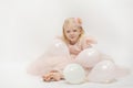 Smiling blonde girl in pink dress and hoop on white background. Isolate. Mischievous child sits on floor in balloons