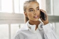 Smiling blonde girl is talking on smartphone. Business woman in white shirt, headphones use mobile phone to work Royalty Free Stock Photo