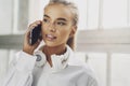 Smiling blonde girl is talking on smartphone. Business woman in white shirt, headphones use mobile phone to work Royalty Free Stock Photo