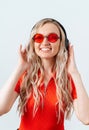 Smiling blonde holds headphones on her head with her hands .. Girl in a red T-shirt and glasses on a white background. Royalty Free Stock Photo