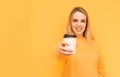 Smiling blonde in an orange sweater shows a cup of coffee in the camera on a yellow background, looking into the camera.Ppaper cup Royalty Free Stock Photo