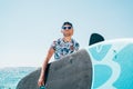 Smiling blonde surfer man in blue sunglasses and cap carrying the surfing board on the bright sunny day noon. Active people summer