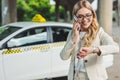 smiling blonde woman in eyeglasses talking by smartphone and checking wristwatch while standing near taxi cab Royalty Free Stock Photo