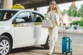 smiling blonde woman in eyeglasses talking by smartphone and looking away while standing with luggage near taxi cab Royalty Free Stock Photo