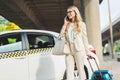 smiling blonde woman in eyeglasses talking by smartphone and looking away while standing with suitcase near taxi cab Royalty Free Stock Photo