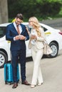 smiling blonde woman holding coffee to go and looking at handsome man using smartphone while standing with suitcase Royalty Free Stock Photo