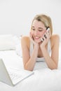 Smiling blonde woman lying on her bed phoning using her notebook Royalty Free Stock Photo
