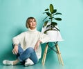 Smiling blonde woman in sweater, jeans and sneakers sits on floor at modern chair with ficus in pot on it looking up