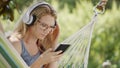 Smiling blonde woman wearing glasses using smartphone, listening to music with headphones, on hammock in the garden, leisure and Royalty Free Stock Photo