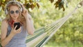 Smiling blonde woman wearing sunglasses using smartphone, listening to music with headphones, on hammock in the garden, leisure Royalty Free Stock Photo