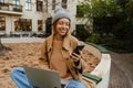 Smiling blonde woman working on laptop and using smartphone while sitting in yard