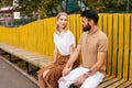 Smiling blonde young woman and confident bearded man with tattooed hand having carefree talk sitting on bench holding Royalty Free Stock Photo