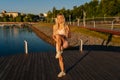 Smiling blonde young woman preparing for cardio workout, stretching leg muscles, holding knee to chest standing by water