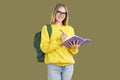 Smiling blondy student girl in glasses with backpack makes notes in copybook on khaki background.