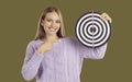 Smiling blondy woman holding dartboard shooting target and pointing finger on khaki background.