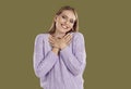 Smiling blondy woman in purple sweater keeping hands at chest or heart on khaki background.