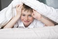 Smiling boy hiding in bed under a white blanket or coverlet. Royalty Free Stock Photo