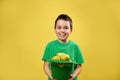 Smiling boy holds a leprechaun green Irish hat full of apples and smiles at camera. Saint Patrick`s Day. Yellow Background