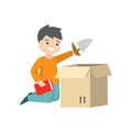 A boy is sitting on the floor and packing his toy sail-boat and a red book in a cardboard box