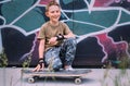 Smiling boy with skateboard sits near grafity painted wall