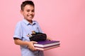 A smiling boy, student at school, holds books and pencil case in front of him. Back to school concepts on pink background with