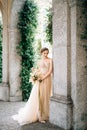Smiling bride in a dress with a bouquet of flowers stands near the arch of an ancient villa. Lake Como, Italy