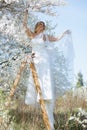 Smiling bride in nuptial dress stand on stepladder under white sakura, hold gauzy fabric of gown while looking away. Royalty Free Stock Photo