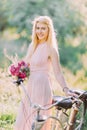 The smiling bridesmaid with the bouquet of the pink flowers is standing near the bicycle in the sunny forest.