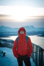 Smiling brunette hidden in a red jacket stands on top of a fjellheisen in Tromso, northern Norway during the Scandinavian winter.