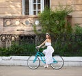 Smiling brunette in white dress riding vintage blue bike down green old street. Royalty Free Stock Photo