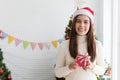 Smiling brunette woman in sweater and SantaÃ¢â¬â¢s hat holding small gift box and rejoices over Christmas decorative Royalty Free Stock Photo