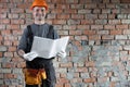 smiling builder with a plan or drawing in his hands standing near a brick wall in the house. An adult engineer in a