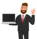 Smiling business man holding or showing a new brand, blank screen laptop computer PC and gesturing, making okay, OK, cool sign