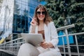 Smiling business woman Graphic Designer in white suit and sunglasses working on digital tablet sitting near modern office building Royalty Free Stock Photo