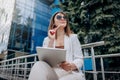 Smiling business woman Graphic Designer in white suit and sunglasses working on digital tablet sitting near modern office building Royalty Free Stock Photo