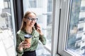 Smiling business woman having coffee break and talking by phone near window at office. Communication, technology, success concept Royalty Free Stock Photo